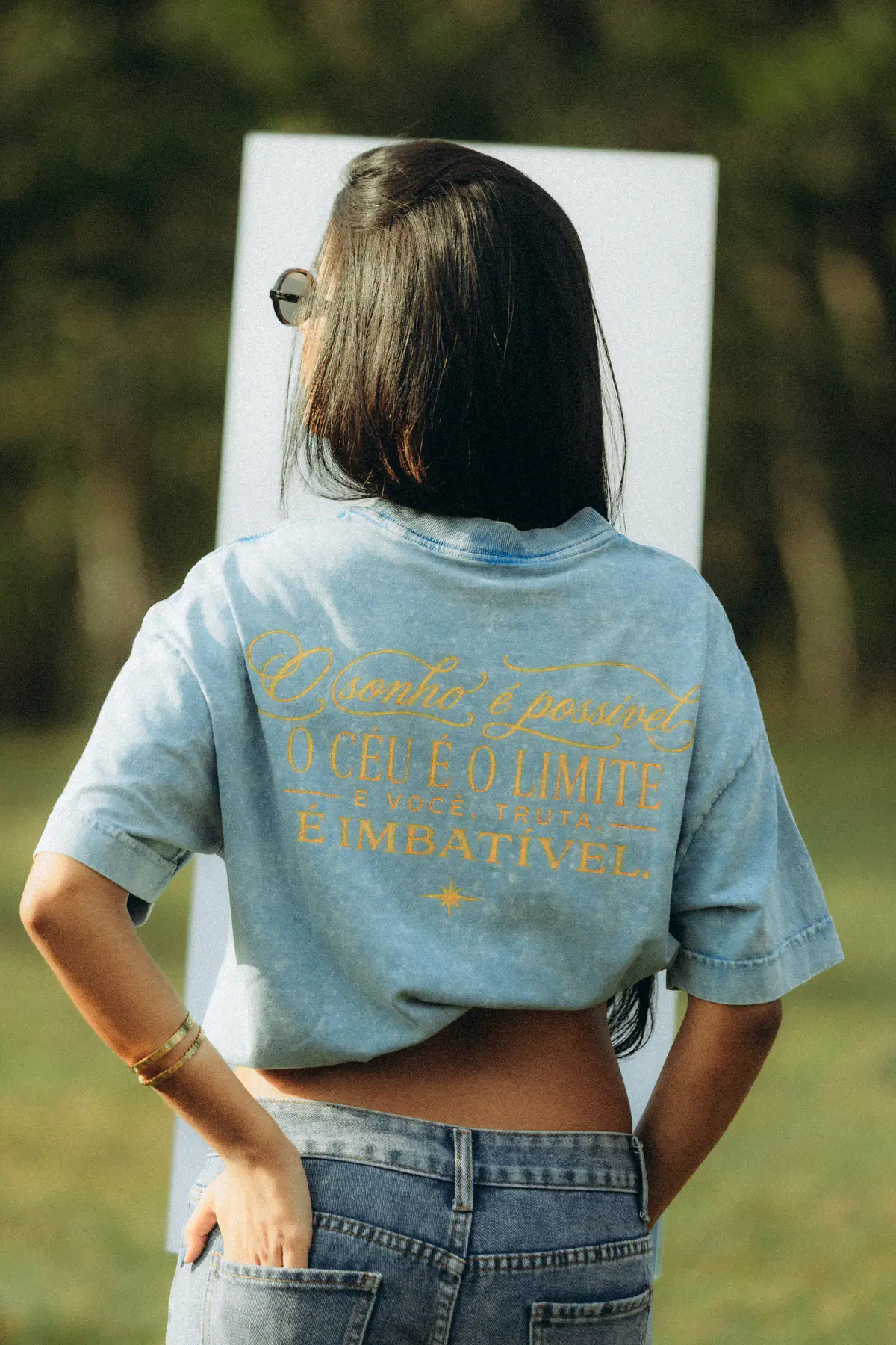 Mulher de costas, de camiseta azul estonada com o texto bordado em dourado: o sonho é possível, o céu é o limite, imbatível.